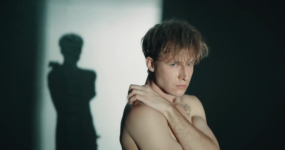 Topless man with tattoo stands in dark room touching his shoulders