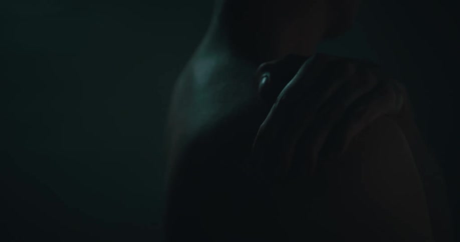 Blonde man holds onto his shoulder as light shines on him in dim area
