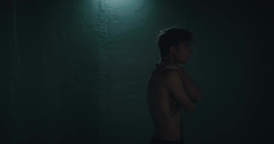 A man without a shirt on and his hands across his chest stands in a dark room with a white light flashing over him