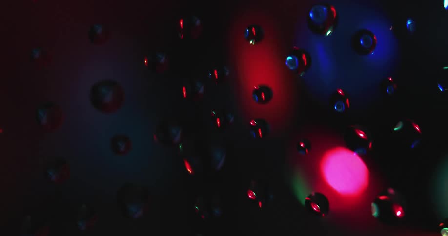 Water droplets sit on clear glass as rotating colored lights move in the distance reflecting light and shadows on the drops
