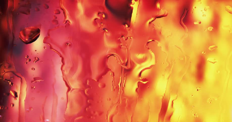 Water droplets drip and move down clear glass as yellow and red light shines and reflects on the water