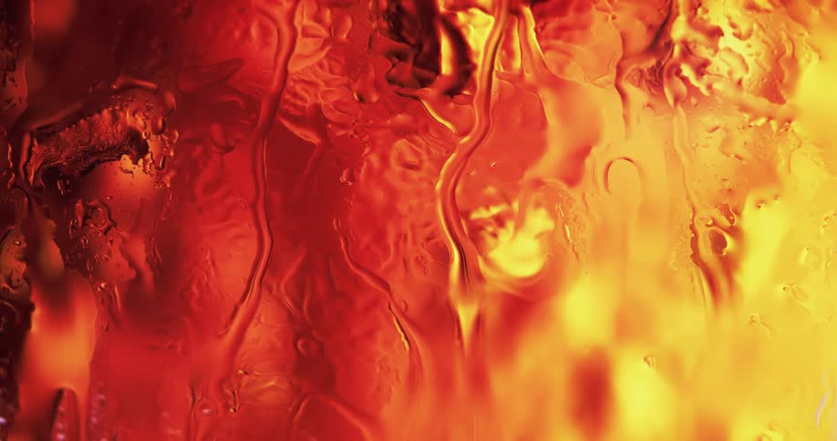 Water droplets drip and move down clear glass as yellow and red light shines and reflects on the moving water