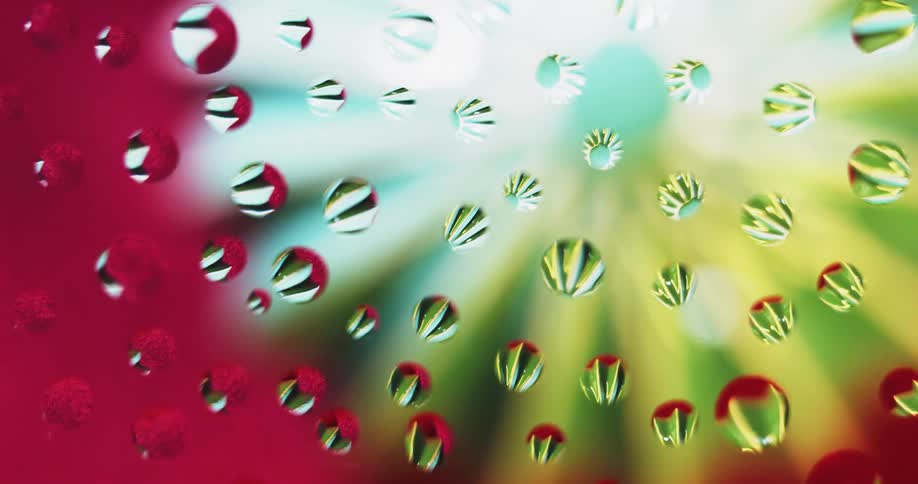 Water droplets are on clear glass as a yellow and pastel blue flower shaped light moves behind them