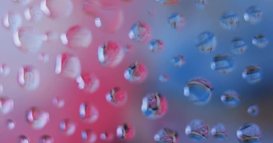 Water droplets sit on clear glass as blue and pink light moves in the distance reflecting on the drops