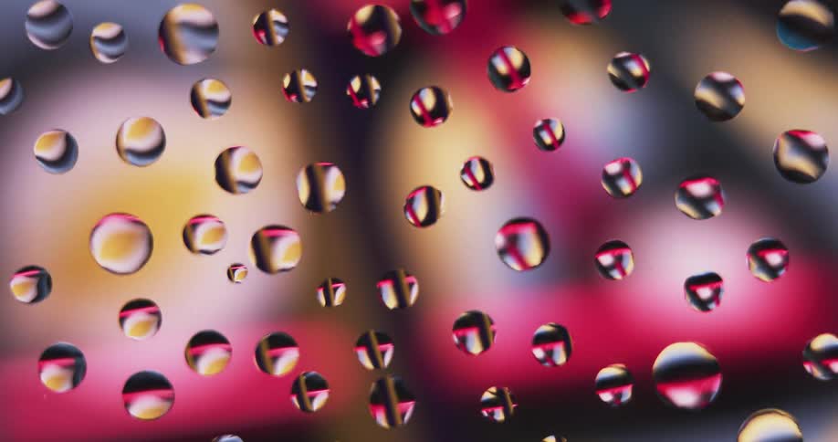 Water droplets sit on clear glass as rotating colored lights move in the distance reflecting on the drops