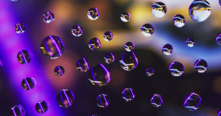 Purple and pink lights move exposing water droplets on clear glass with an amber light in the distance