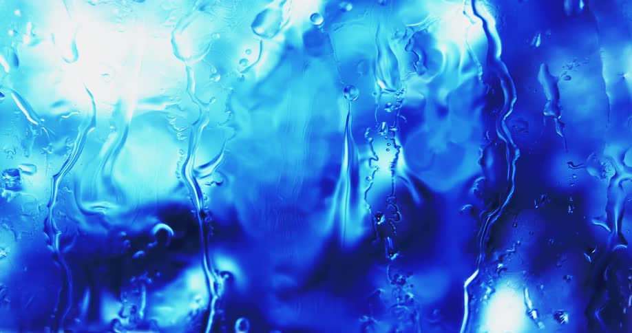 Water droplets drip on clear glass as blue and white light glows in the distance reflecting on the drops