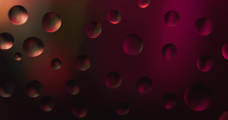 Water droplets sit on clear glass as changing colors of light shine and texture develops inside the drops