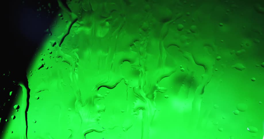 Water droplets fall on clear glass and drip as a glowing green globe shines behind them