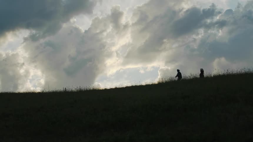Two people are silhouetted against the cloudy sky while walking across top of grassy hill