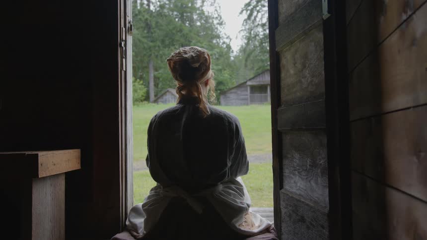 A woman sitting on the doorstep with her back to the camera looking at the yard a shed trees and grass in the background