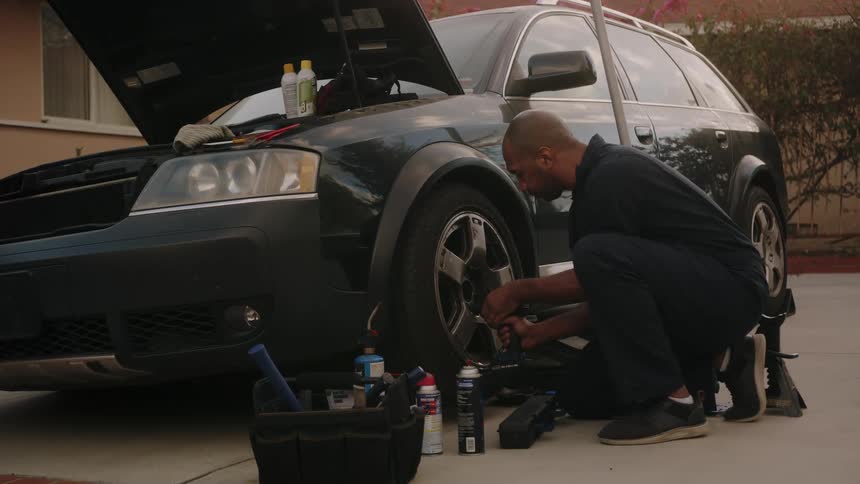 A black man is tightening the lug nuts on a black sedan with a cordless drill