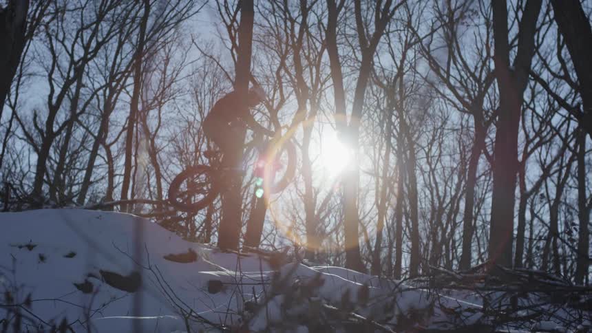 A male jumps down a snow covered hill on his bicycle surrounded by trees