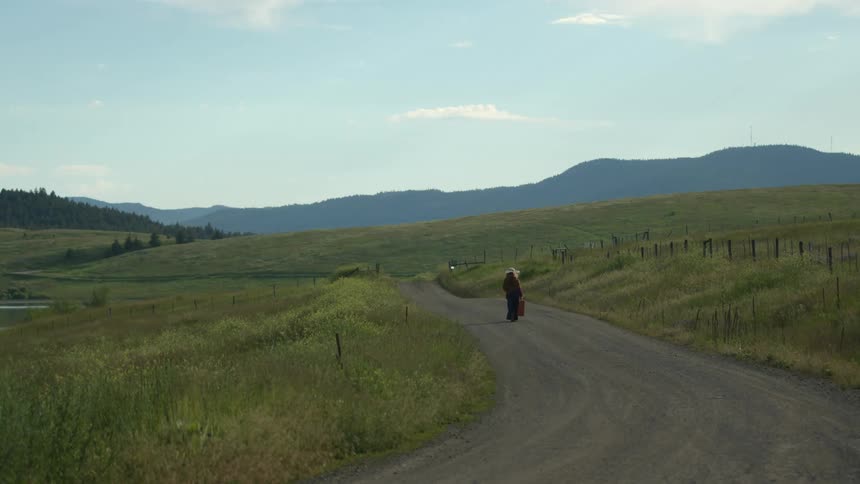 A woman is carrying a shopping bag as she walks on a rural country road with mountains in the distance