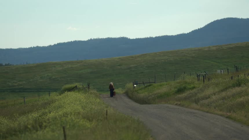 A woman is carrying a shopping bag as she walks on a rural country road with mountains in the distance