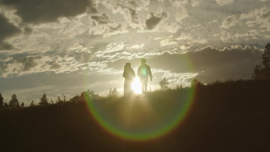 Silhouette of a man and a woman walking on a hill as the sun shines brightly on the horizon under a cloudy sky