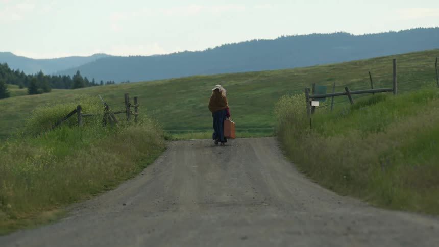 Woman wearing a hat and a long skirt walks with a briefcase on a gravel road in the countryside