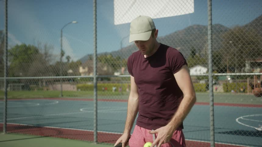 Man bounces tennis ball as he walks forward on tennis court at park near mountains