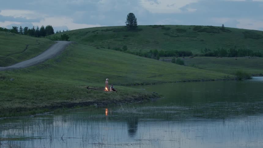 Person approaches campfire and joins person there on riverbank