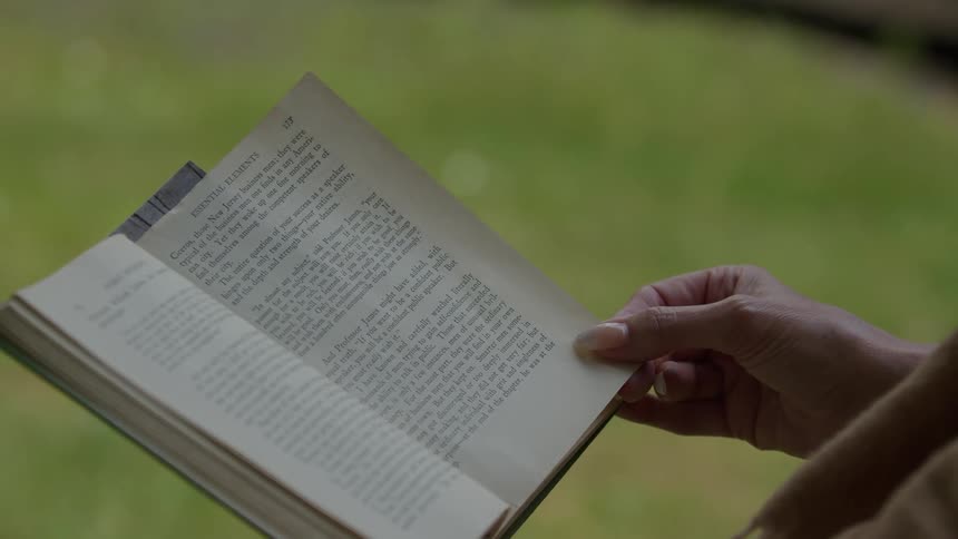 A woman is sitting outside holding a book as the page is moving in the breeze