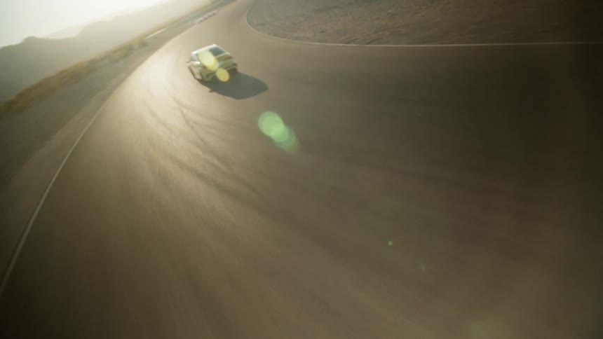 A yellow automobile slides as it speeds around a curve across a desert road with mountains in the distance