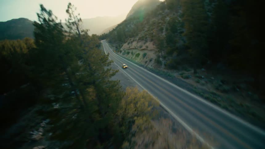 A yellow car driving down the street is filmed by a drone speeding down and over the car
