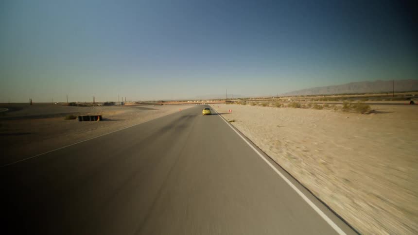 A yellow car is speeding on a desert road with mountains in the distance under a clear blue sky