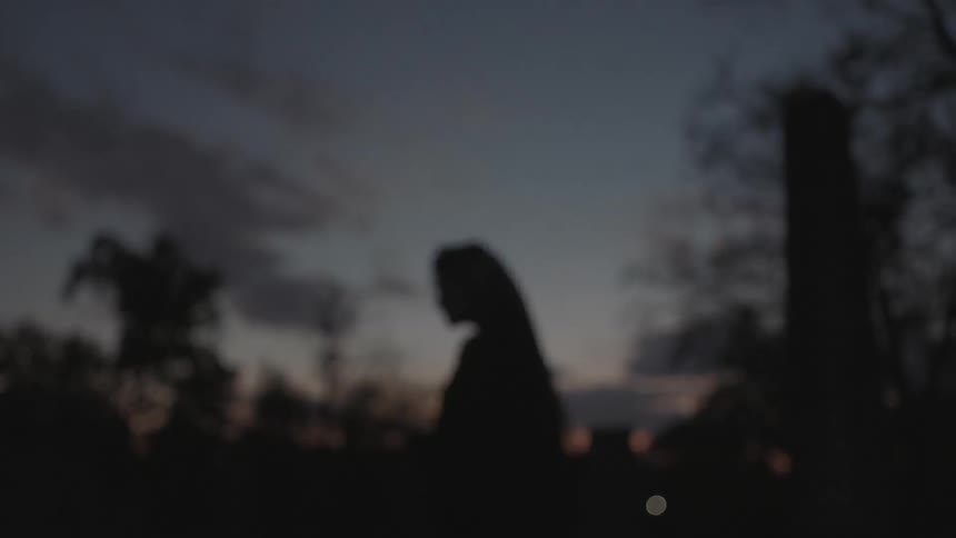A woman in a veil stands in a cemetery at dusk