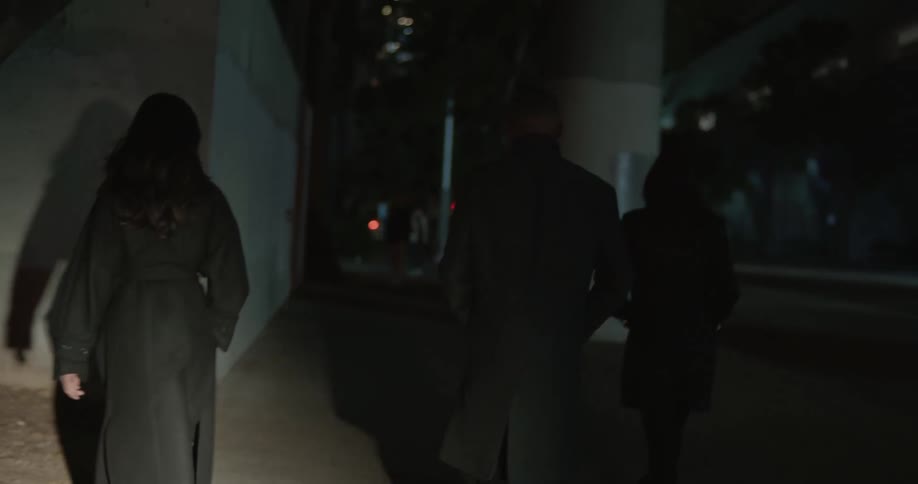 Man in black overcoat and two women wearing black overcoats walk down the street at night