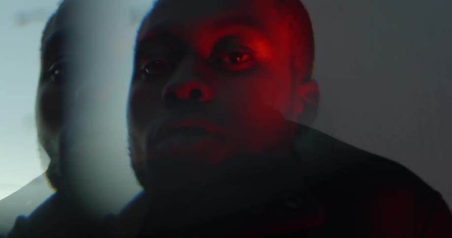 Close up of a man standing next to a window looking at the camera while a red light shines on his face and the reflection of the sky at dusk and a streetlight can be seen in the window next to him