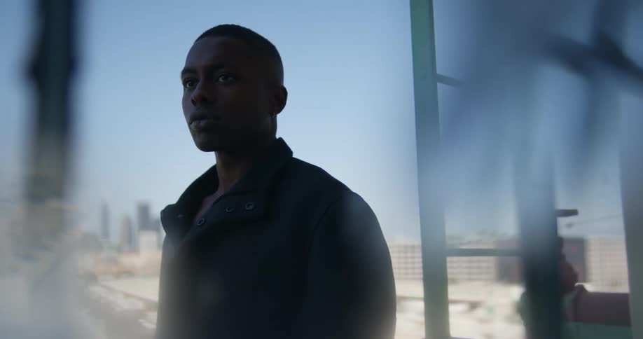 Man with black overcoat stands on top of building with city skyline in the background