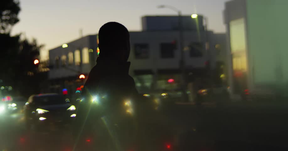 Black man pacing on city side walk at sunset as traffic lights rush by view through glass with reflections from cars