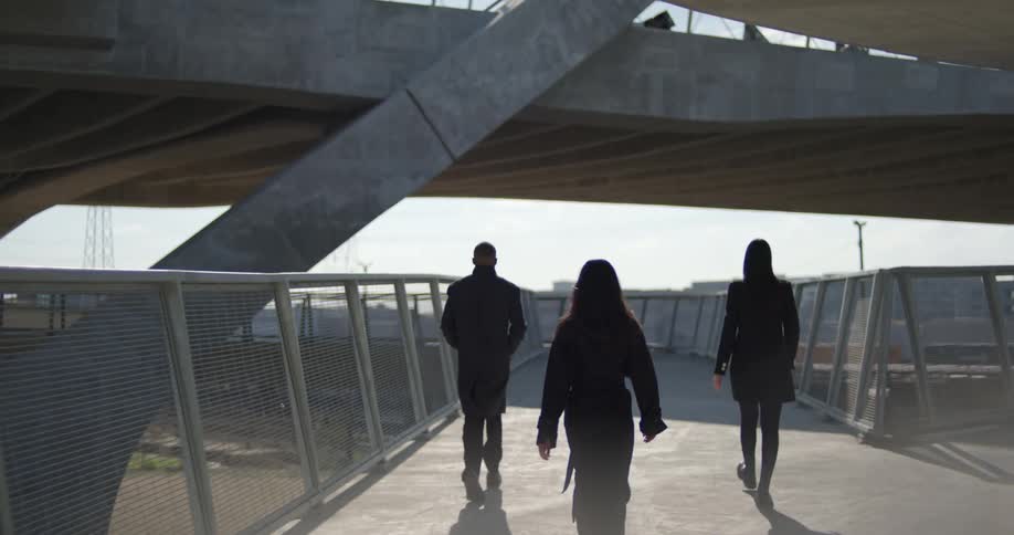Three people move along raised fenced walkway under overpass as train passes on ground level