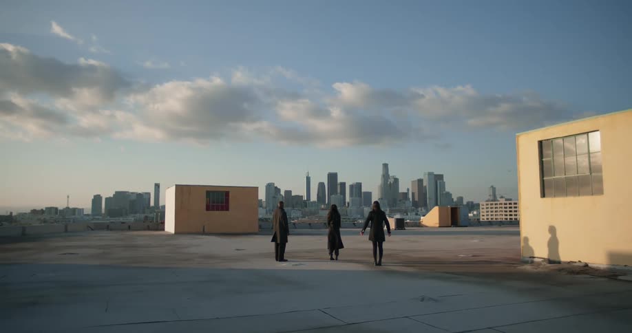 Two woman and a man all dressed in black stand in front of a city scene with many buildings and the blue sky overhead