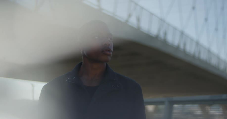 Man with earrings and black overcoat looks around near bridge with city skyline in the background