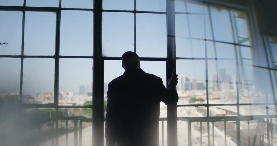 Man with black overcoat and pants looks out the large windows of a building