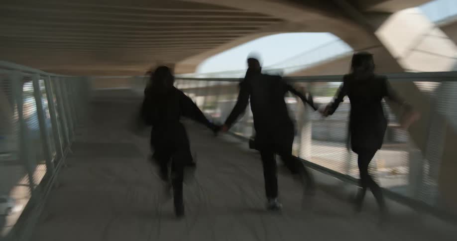 Three people run along footbridge under overpass near train tracks as train passes