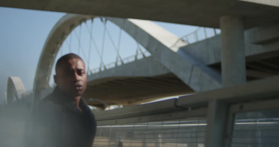 Man wearing coat leans against meshed fence then turns and walks along side it with structure in the background