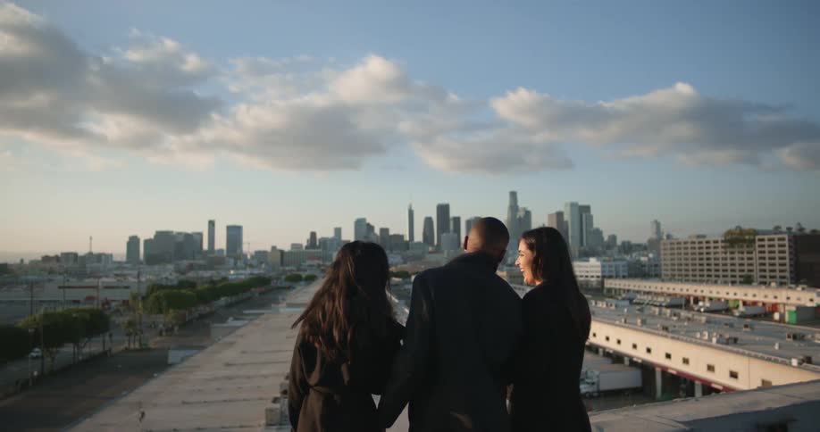 Three friends two white women and one black man all three dressed in black trench coats congregating on a rooftop in the city as camera pushes past them and reveals the city skyline