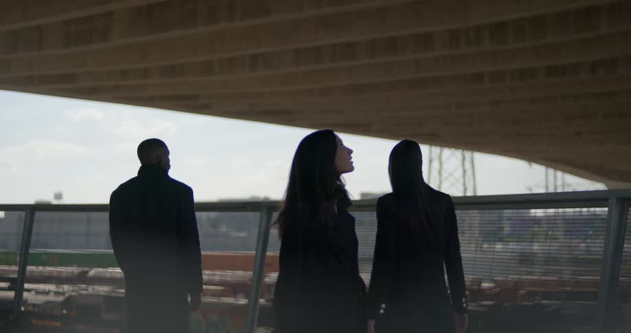 Three people walk along fence enclosed path under overpass near train tracks