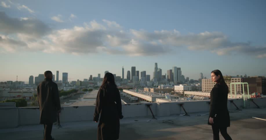 Man in black overcoat shows city skyline to two women wearing black overcoats pants and shoes on top of building