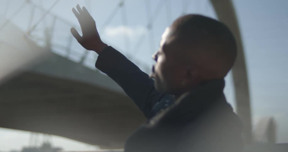 Man holds up hand to block sun while looking toward bridge connected on one end to city downtown