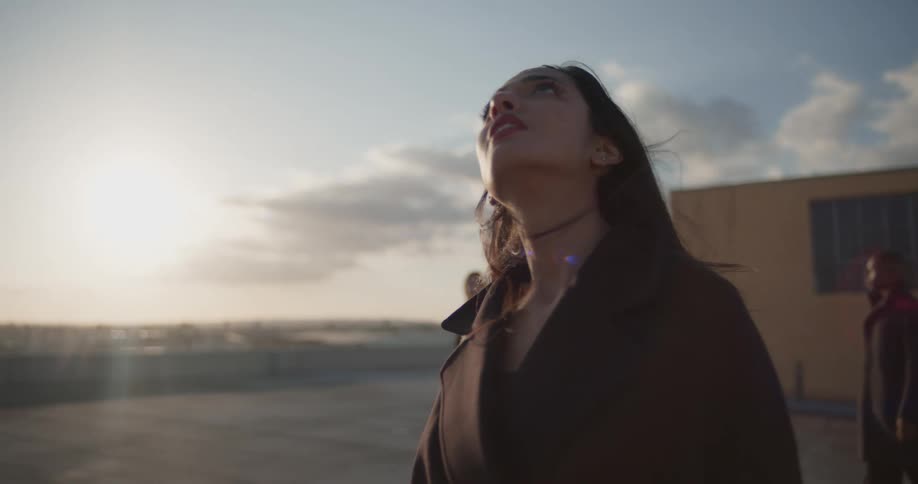 Man in black coat and women wearing black coat and blazer jacket walk and look up on top of building on a sunny day