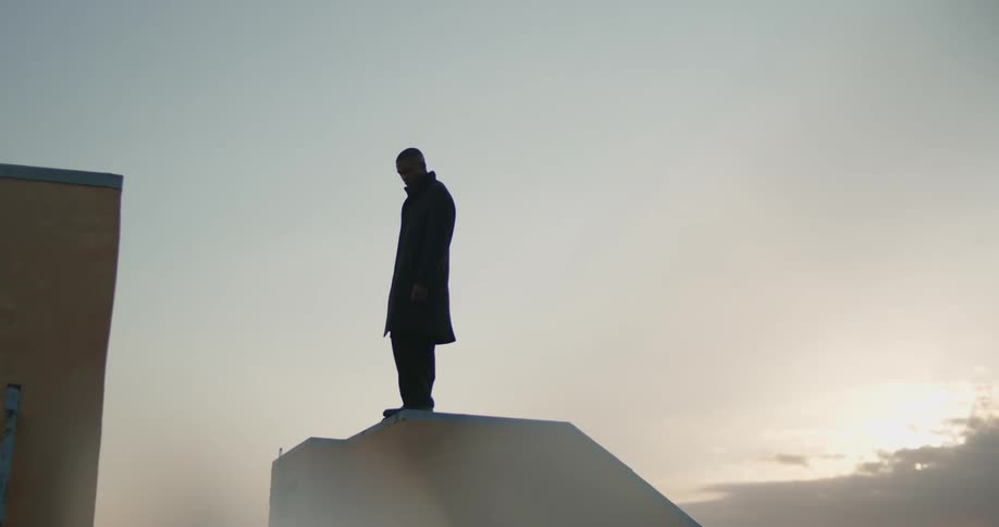 A man dressed in black stands high up on the edge of something and puts one foot out as if hes going to jump off