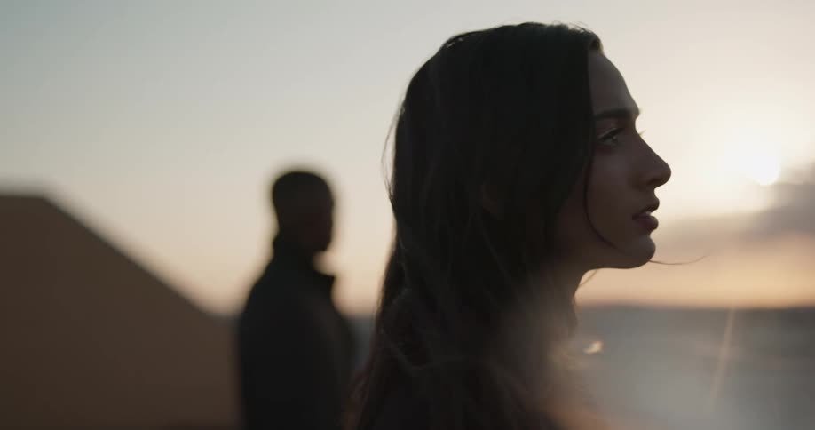 Man in black coat and women with dark hair and black coat are silhouetted against the sky at sunset on top of building watching city skyline