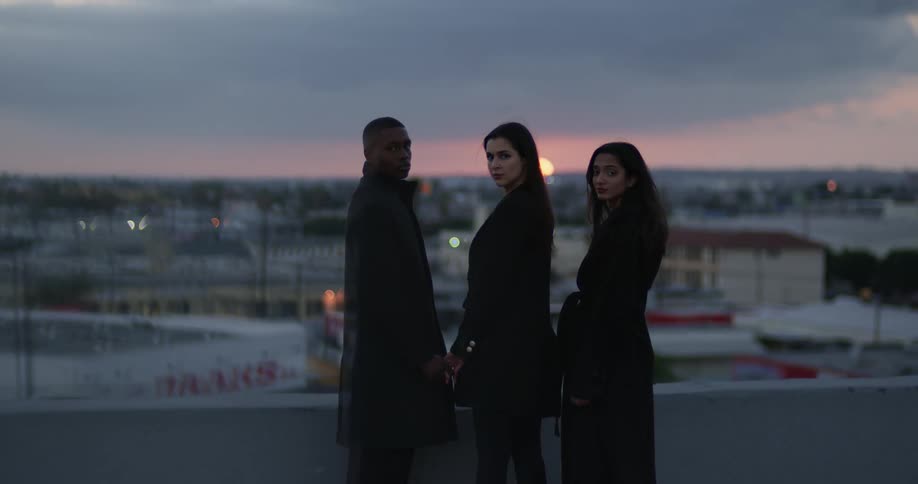 Man in black coat and women wearing black coat and blazer jacket stares at camera on top of the building at sunset