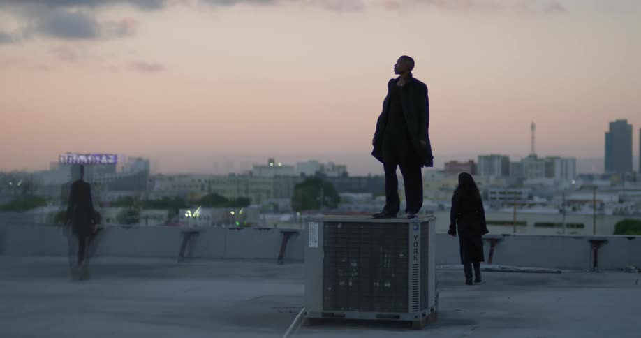 Man stands on ac unit as his friends move across building roof with skyline in distance