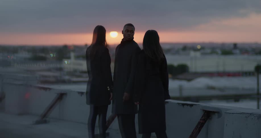 Man stands between two women looking toward one as sun sits in red sky over city