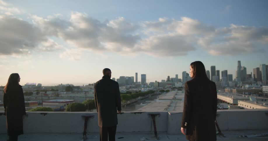 Man in black coat and women wearing black coat and blazer jacket watch city skyline on top of building