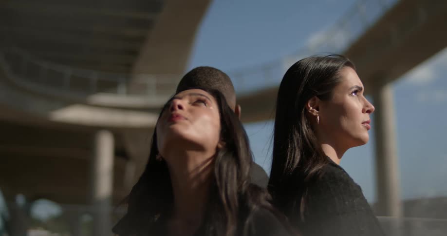 Two white women and one black man standing back to back in a threesome under and underpass looking out into the world as the camera orbits around them
